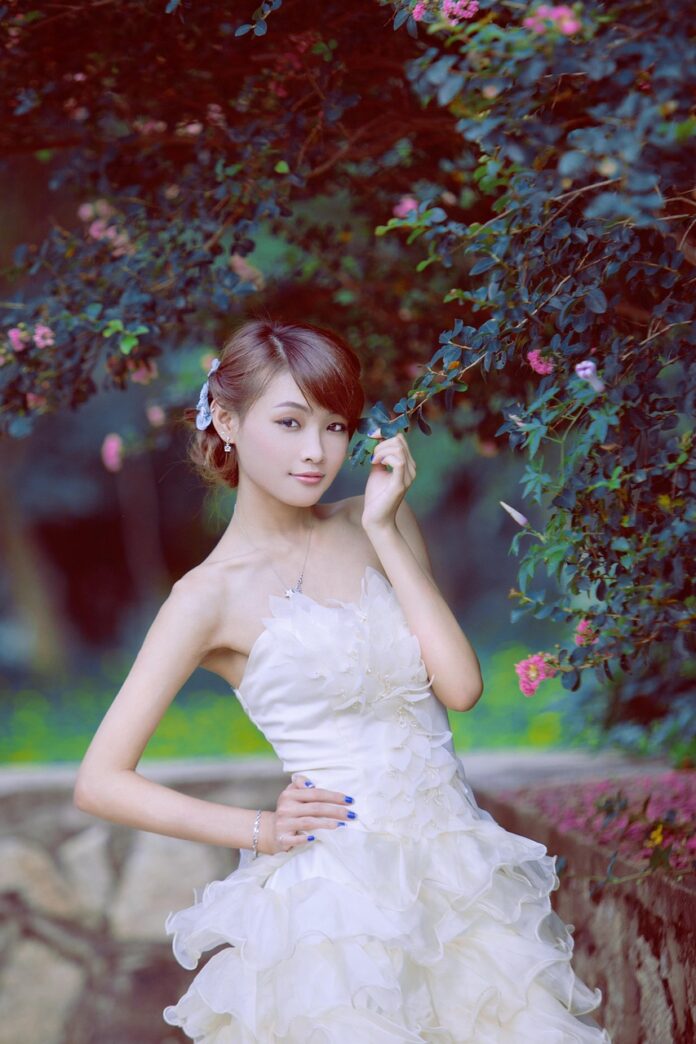 Bride in a strapless white, petal-embellished wedding gown posing beside pink flowering bushes in a garden.