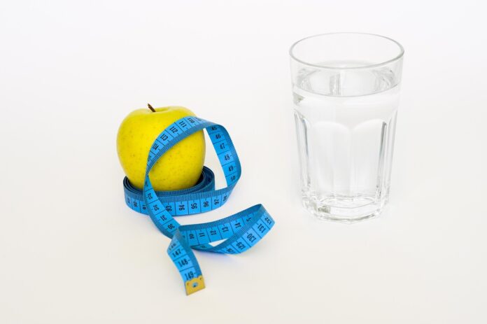 Yellow apple with a blue measuring tape wrapped around it, next to a glass of water on a white background — health and diet theme