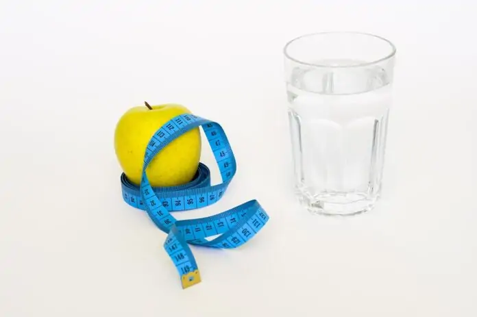 Yellow apple with a blue measuring tape wrapped around it, next to a glass of water on a white background — health and diet theme