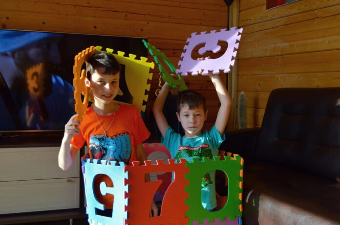 Two kids stand inside a bright, multicolor foam block play structure with interlocking letter panels in a wood-paneled room.
